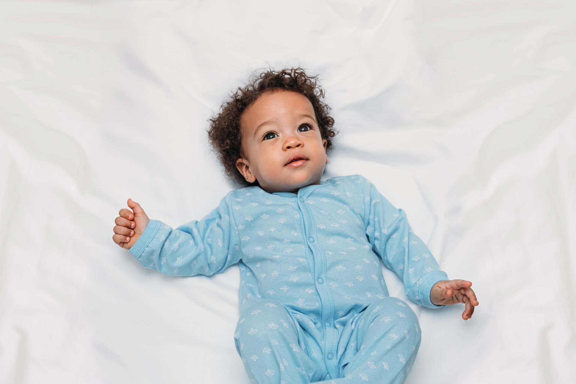 Baby wearing a blue pajama laying in a bed with white sheets.